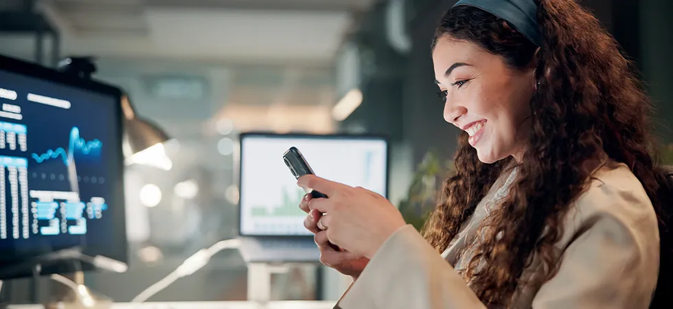 A smiling professional using a smartphone to monitor real-time network data dashboards for MNO customer support outsourcing.
