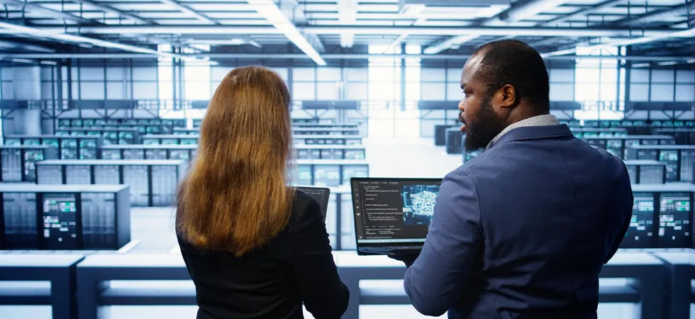 Data center access governance coordinators reviewing facility entry logs on a laptop inside a server hall